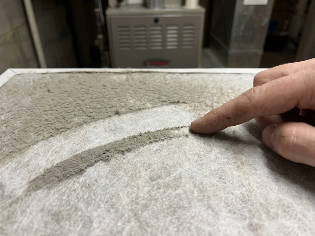 A hand swiping a finger across an HVAC filter showing a gray dust streak.