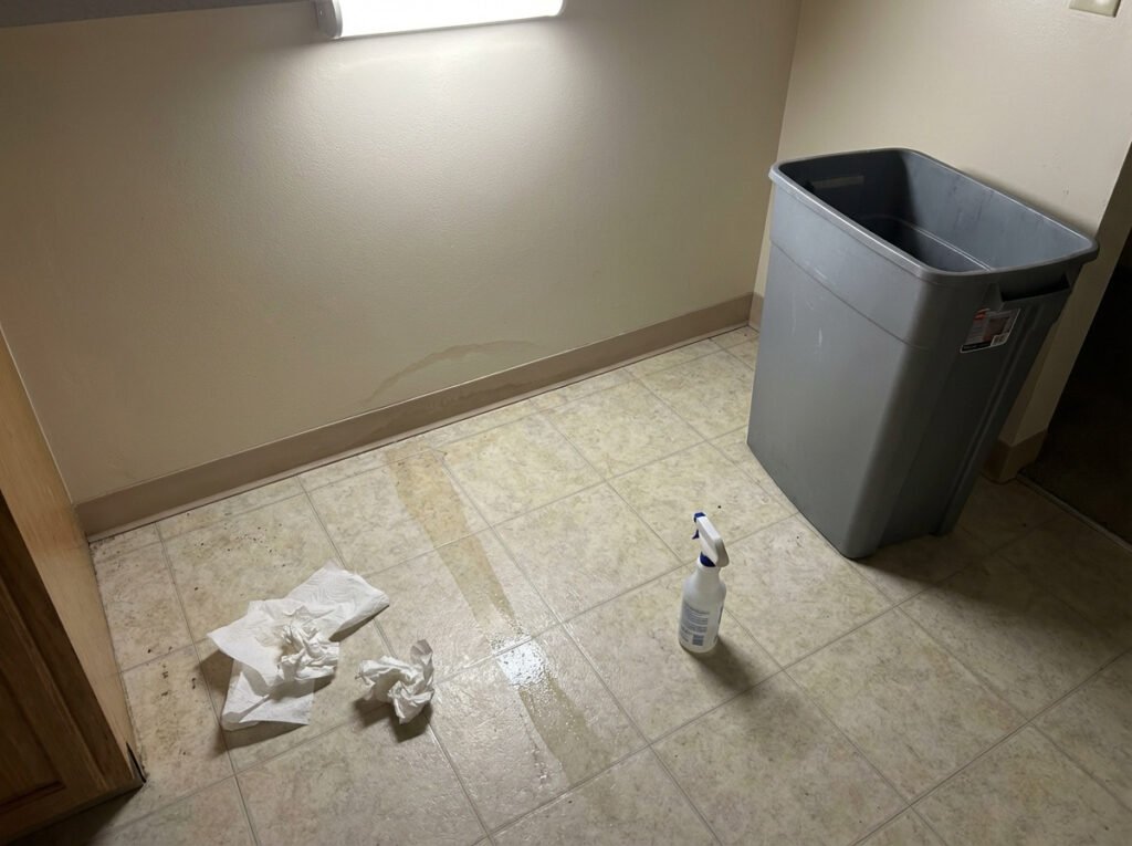 A trash can pulled away from the wall showing a cleaned area on the floor where a leak trail was found.